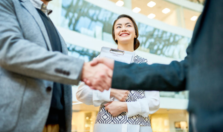 Pretty assistant manager with clipboard in hands looking at entrepreneurs with wide smile while they shaking hands after successful completion of negotiations